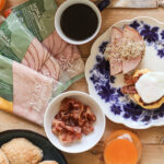 En vit tallrik med blått mönster med eggs Benedict och alfalfagroddar, en skål med knaperstekt bacon och tre paket smörgåsskinka i Smak av Sveas gröna förpackning. Omgärdade av kaffe, juice övrig frukost.