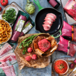 Köttprodukter i och utanför förpackning ligger upplagda på fat och skärbrädor omgärdade av grönsaker och spannmål. Fotograferat ovanifrån.